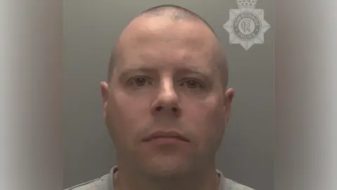 A police custody shot of Ian Walpole. He has a bald head and is looking at the camera. He is wearing a light grey t-shirt. The wall behind him is grey.