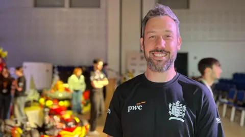 Carl is wearing a black PwC T‑shirt stands in a hall with a brightly coloured go‑kart display and several people in the background