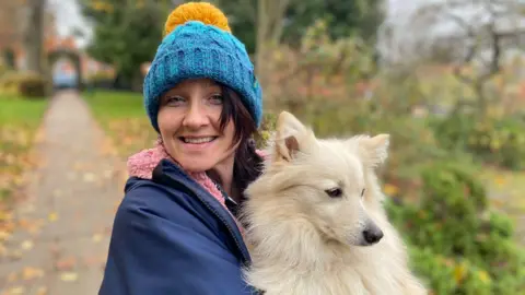 Hannah Perkins, standing in a park, looking at the camera, wearing a warm coat and bobble hat, holding a small white dog.