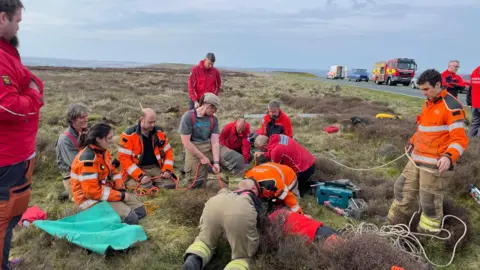 Scarborough and Ryedale Mountain Rescue Team A group of people in high-visability outfits kneel and stands around an area of moorland. They hold ropes and other rescue equipment.