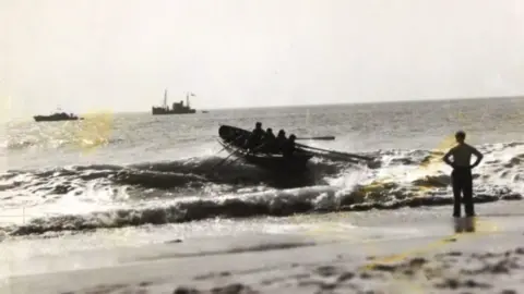 Falmouth Maritime Muesum A black and white wartime picture of a rowing boat landing on a beach in the surf. In the background is a fishing vessel and one of the gunboats that towed it ashore. 