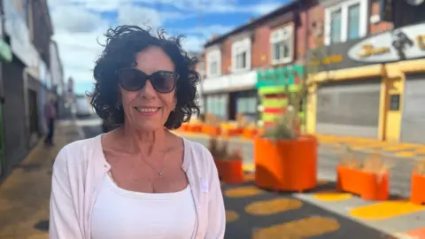 A woman with black hair and sunglasses stands on a street, smiling at the camera. In the background you can see brightly coloured paint on the street and orange planters. 