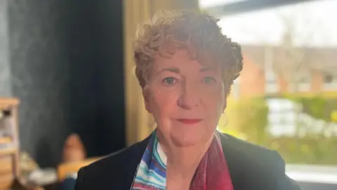 A woman with short, curly blonde hair looks into the camera. She is wearing a dark jacket and a multi-coloured scarf. A window is in the background.
