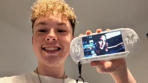BBC Kyle is indoors holding up a white PlayStation Portable (PSP) console with a keychain attached.