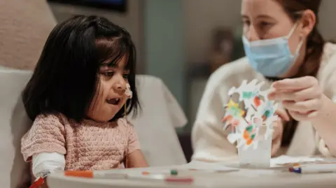 Zarwa is pictured sitting at a hospital table smiling as a masked staff member helps her make a colourful paper snowflake during an arts and crafts activity.