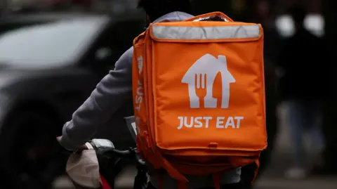 Reuters A person photographed from behind, cycling, wearing an orange and white Just Eat backpack and holding a white cap