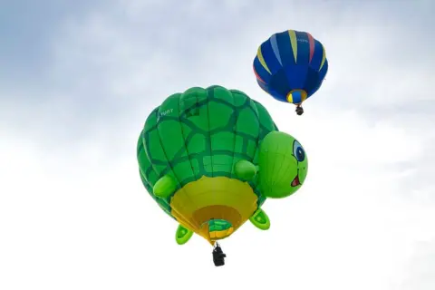 Claire Hartley A hot air balloon shaped like a green turtle is in the sky, just behind is a blue balloon.