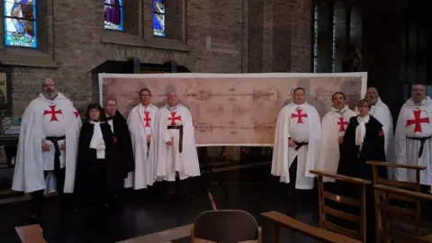 A group of people, some dressed in white robes with red crosses, stand inside a church in front of a large replica of the Shroud of Turin. The church features stained-glass windows and wooden chairs.