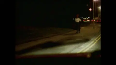 A woman in a white top with dark skirt is seen arguing with a man at night by the side of the road in a BBC reconstruction