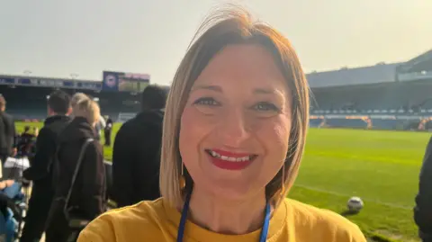 Gemma Nichols has blonde hair and is wearing a mustard top and a blue lanyard. In the background a football pitch and a football is visible.