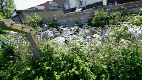 Sureena Brackenridge A gated off area between allotments and a row of houses, showing mounds of rubbish piling up.
