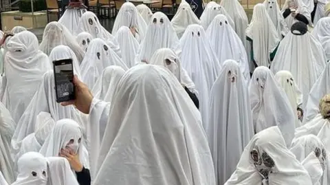 A group of people with white bed sheets over them - appearing like ghosts.