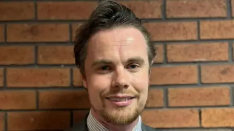 George King/BBC The head and shoulders of Phillip Faircloth- Mutton in front of a brick wall. He appears to be wearing a striped shirt. He has a large gelled quiff and a trimmed beard.