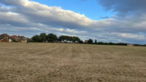 wide, open grass field with dry, brown turf stretches across the foreground under a dramatic sky filled with large grey and white clouds. A line of trees, small football goals, and houses are visible along the horizon, with a few distant people walking near the left side of the field. Patches of blue sky break through the cloud cover, casting soft light across the landscape.