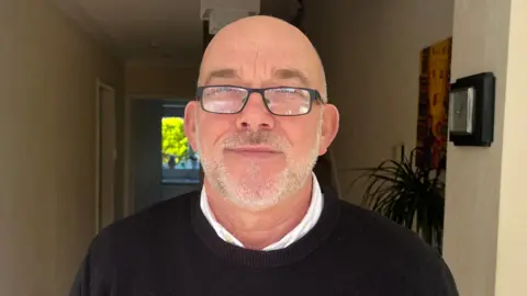 A man with balding grey hair and a beard and wearing a white shirt and dark coloured jumper is smiling as he stands inside the hallway of a house.