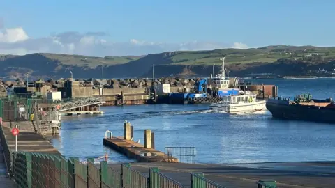 BBC A small harbour scene shows a white passenger boat moving through calm water toward a quay, with a larger blue vessel moored nearby. The harbour is lined with metal railings, concrete walkways and a ramp leading down to the water. Beyond the breakwater, rolling green hills rise in the distance under a bright blue sky.
