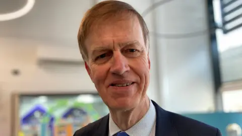 A man in his sixties with light brown hair wearing a smart shirt, tie and blue jakcet in the library of Treloar College