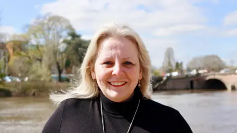 Elena Round pictured wearing a black turtle necked jumper with a silver chain. She has long wavy blonde hair. 