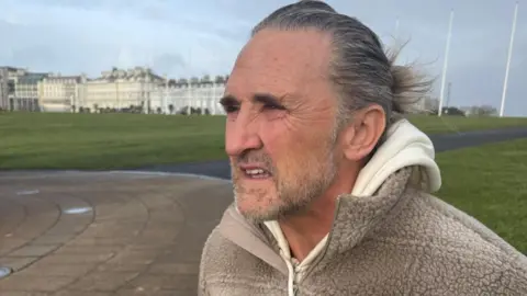 Paul Maxwell, wearing a beige fleece and hoodie, looks towards the left of frame. He is sitting on a bench on Plymouth Hoe.