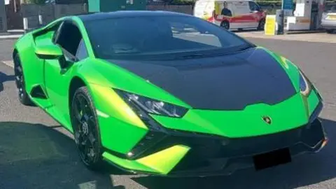 A green Lamborghini, parked up in a petrol station. It is green with black bits on its hood. A van is in the distance.