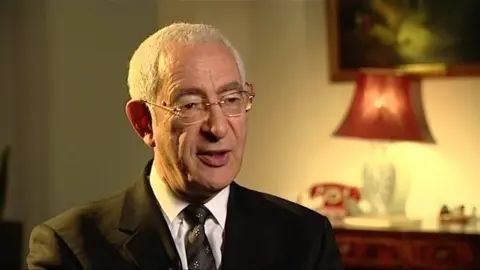 BBC Lord David Triesman, wearing a black suit and speaking to the camera, in the dimmed light coming from a red lampshade 