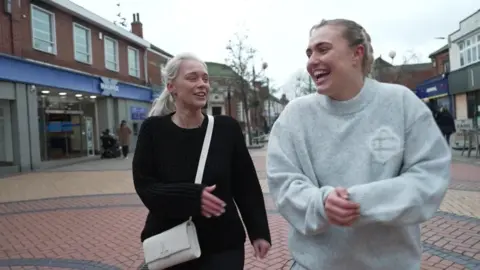 BBC / Matt Dean Faith Anderson is on the left of the image and is looking at Gemma and smiling, she is wearing a black jumper and a cream cross-body bag. 
Gemma is wearing a grey jumper and is laughing. The women are walking together down a pedestrian shopping street.
