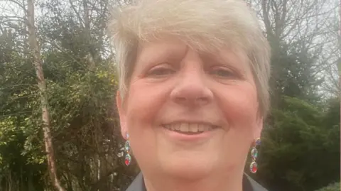 Supplied A photograph of a woman with short hear wearing hanging earrings. The photograph is taken outside and trees can be seen behind her.