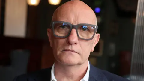 PA Colin Neill has thick-rimmed black glasses. He is wearing a black suit with a white shirt. The background behind him is blurred. 