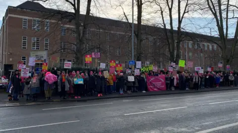 A line of people holding unreadable placards standing on a path outside a large red brick building.