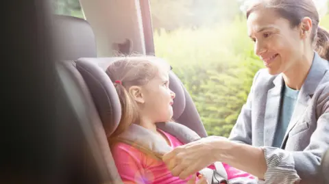 A woman putting a little girl in a car seat. The woman is wearing a grey jacket and blue jumper. She has her hair tied back and is smiling. The little girl is looking at her and wearing a pink top.