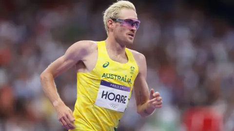 Man with bleached blonde hair and wraparound sunglasses running. He is wearing a bright yellow Australia vest.