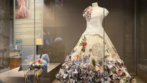 A white dress with colourful flowers sewn onto the skirt and left shoulder is on display in a glass case in a museum. 