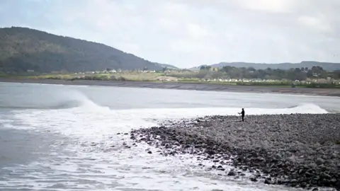 Getty Images View of Pollock Weir. Waves are coming in as a surfer in the distance is getting ready. Porlock is in the background in front of a hill.