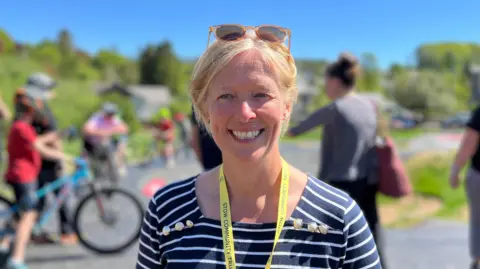 A blonde haired woman with sunglasses on her head and blue striped top smiling on camera