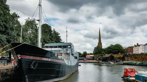 DHP Family A cargo ship bearing the name "Thekla" is moored on a city's harbourside on a cloudy day. Around it, other smaller boats are using the harbourside while further away a brown, beige and light blue building can be seen, as well as a church spire.