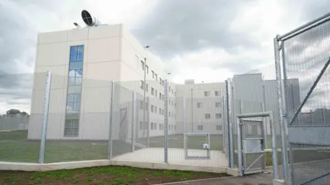 PA Media An exterior shot of HMP Fosse Way. A white building is protected by a metal fence. On the roof is a fan.