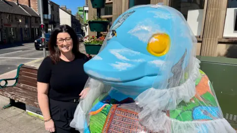 A giant duck sculpture sits outside a former bank building with a white wedding veil on its head. The duck has a detailed drawing of badgers, a fox and birds over a countryside view of a mill beside a river. It's eyes are yellow. A woman stands next to the sculpture win a black t-shirt, black trousers, black glasses with black hair. There is a wooden bench, flower pots, a set of traffic lights and a car in the background.
