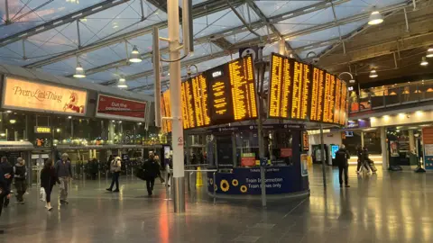 Photograph of the concourse at Manchester Piccadilly Station. The image shows the departure board and passengers making their way to the platforms.