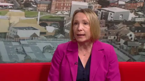 Helen Morgan in a pink jacket over a black top. She is sitting on a red sofa in the TV studio
