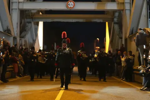 PA Serving paratroopers from the Parachute Regiment, the Rifles, the Army Air Corps and the Salamanca Band and Bugles of the Rifles, march at the double across Pegasus Bridge