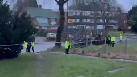 @num1dad91 Police officers have taped off the scene of an incident where a tree has fallen on a car. There is a grass area in the foreground, and a block of flats in the distance.
