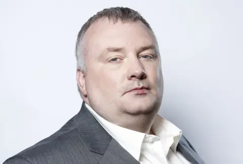 A man with short hair is turned to the side looking into the camera. He is wearing a white shirt and grey blazer.