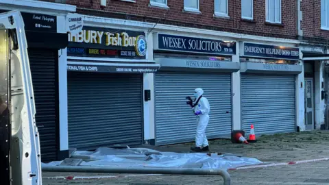 A member of a forensic team with a white suit taking photographs in the area of the stabbing with a camera. Two cones lay nearby. A door of a white van is seen open in the picture frame.