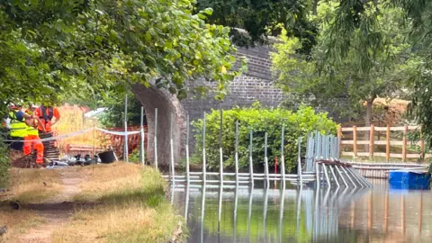 A section of canal running under a bridge, with workers seen standing on the towpath wearing orange high visibility clothing. There is a metal structure in the water, which appears to be the framework of a temporary dam.