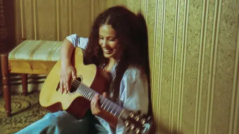 Black Butter Records Jessy is sat on the floor, her back against a brown-wallpapered bedroom wall. She is smiling and playing an acoustic guitar.