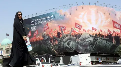 A woman clad in black walks in front of a poster that shows a toppled Statue of Liberty in front of a crowd waving red flags