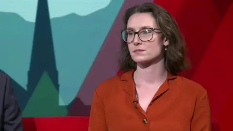 Beth wears glasses and her hair is cut into a bob. She wears a red buttoned top and is sat in the studio on a red sofa.