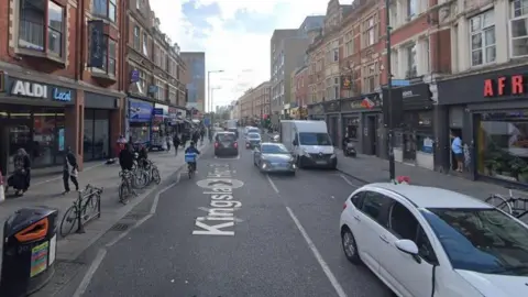 A Google street view screengram of Kingsland High Street with cars and vans on the road and shops lining the street. 