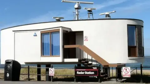 An artist's impression shows how the radar station would look once converted into a flat. It is a single-storey, elliptical-shaped build with windows on its surface and a short staircase leading up to a door. It is on stilts just on the beach behind the promenade railings.