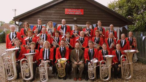 Photograph of band wearing red blazers, black ties and holding their brass instruments